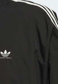 Schwarze Basketballjacke mit weißen Streifenakzenten, aus strukturiertem Stoff und mit gesticktem Logo auf der Brust.