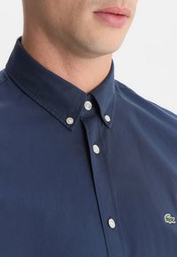 Donkerblauw overhemd met knopen en kraag, voorzien van witte knopen en een klein geborduurd logo op de borst. Gladde textuur.