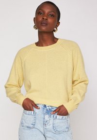 Donna che indossa un maglione giallo chiaro e jeans blu chiaro, con le mani nelle tasche anteriori, orecchini a cerchio dorati, trucco neutro e capelli corti.
