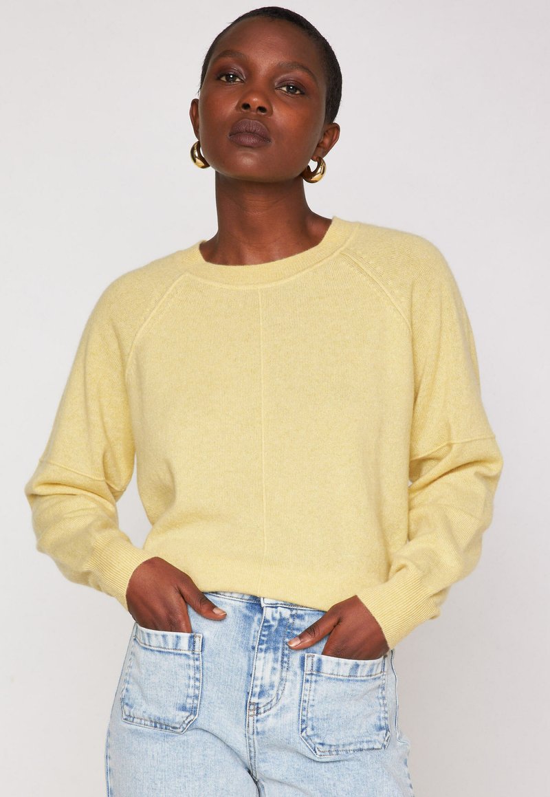 Donna che indossa un maglione giallo chiaro e jeans blu chiaro, con le mani nelle tasche anteriori, orecchini a cerchio dorati, trucco neutro e capelli corti.