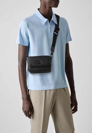 Schwarze gesteppte Umhängetasche mit verstellbarem Riemen, ausgestattet mit einem oberen Deckel und Logodetail. Getragen über einem hellblauen Polo und beigen Hosen.