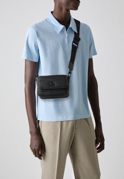 Sac bandoulière noir matelassé avec sangle réglable, doté d'un rabat supérieur et d'un détail de logo. Porté sur un polo bleu clair et un pantalon beige.