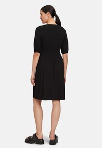Schwarzes Kleid mit kurzen Puffärmeln, tailliertem Schnitt und knielangem Rock. Kombiniert mit klobigen schwarzen Sandalen mit Nietenakzenten.