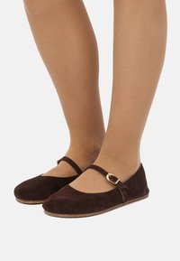 Zapatos planos de ante marrón con una correa y detalle de hebilla, punta redondeada y textura suave, sobre un fondo neutro.