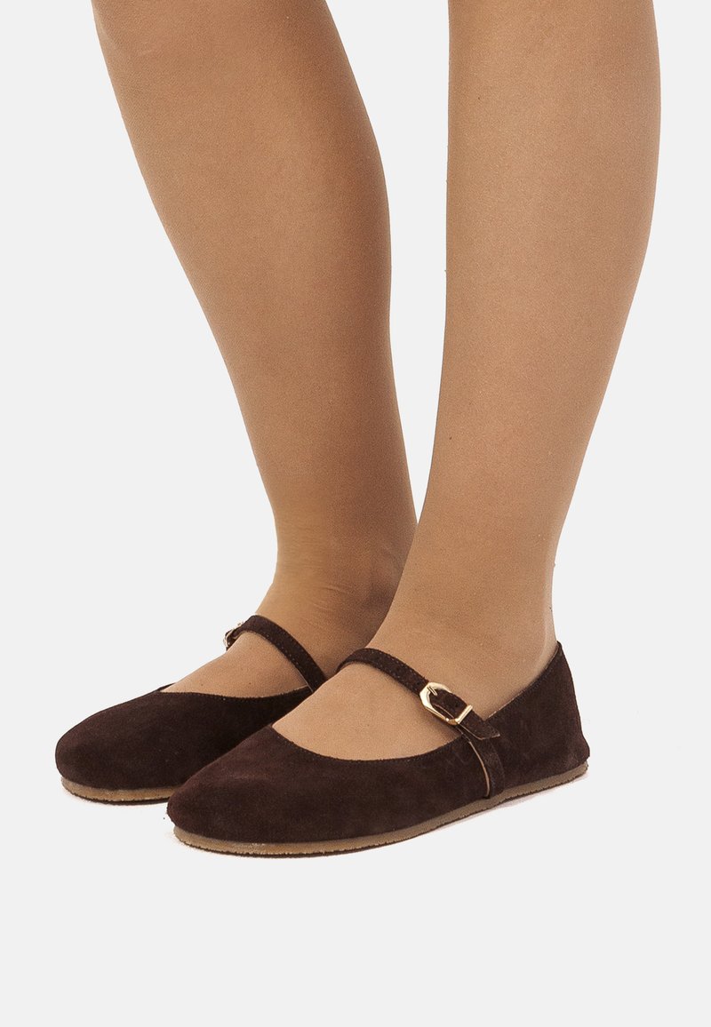 Zapatos planos de ante marrón con una correa y detalle de hebilla, punta redondeada y textura suave, sobre un fondo neutro.