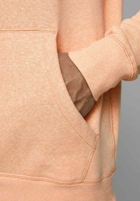 Helle Pfirsich-Pulloverkapuzenjacke mit einer vorderen Tasche. Weicher Stoff, lockere Passform und gerippte Bündchen. Auffällige Nahtdetails und subtile Textur.