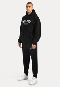 Czarna bluza z kapturem z przednią kieszenią i białym napisem "SMILOOXX ATHL WEAR". W zestawie z czarnymi spodniami z podobnym brandingiem, noszona z białymi trampkami.