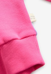 Gaiši rozā džemperis ar ribotām piedurknēm. Redzams mazs auduma birkas uzraksts "Little Bird". Mīksta tekstūra.