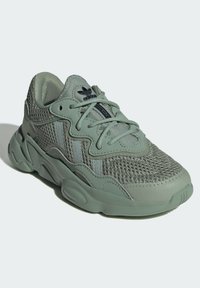 Green athletic shoe with mesh upper, textured accents, rubber sole, and lace-up design featuring three contrasting stripes on the side.