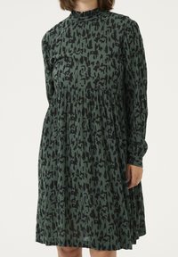 Langärmliges Kleid aus olivgrünem Stoff mit einem schwarzen abstrakten Muster, einem gerafften Ausschnitt und einer gedrapierten Taille für Volumen.