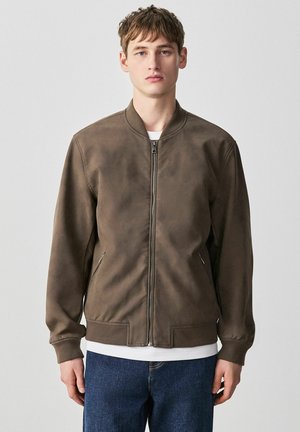 Young man wearing brown zip-up bomber jacket, white shirt underneath, and blue jeans, standing against light gray background.