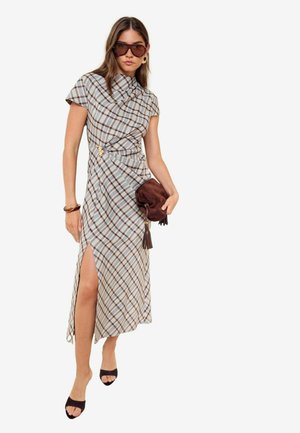 Kvinde iført en ternet midikjole med slidse til låret, der holder en brun clutch, iført solbriller, hoop-øreringe og sorte åbne høje hæle.