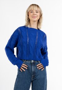 Pulover scurt tricotat de un albastru regal, cu modele din cablu și dantelă, mâneci lungi, manșete cu nervuri și un decolteu rotund. Asortat cu blugi cu talie înaltă.