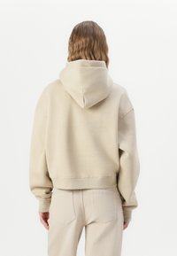 Persona con capelli mossi che indossa un hoodie beige e pantaloni coordinati, in piedi rivolto verso l'esterno su uno sfondo bianco semplice.