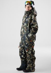 Camouflage sneeuwpak in groene en beige tinten, voorzien van ritszakken, oranje accenten en zwarte sneeuwschoenen. Inclusief goggles en een beanie.