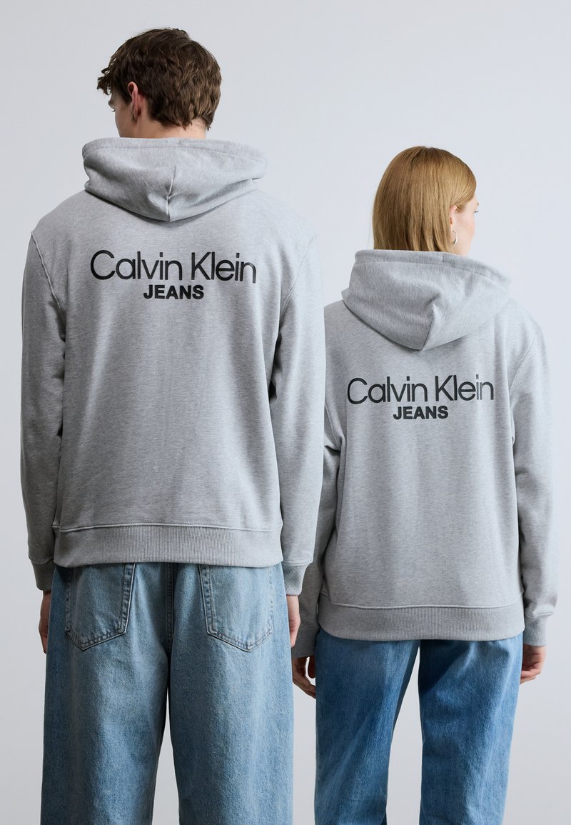 Γκρι φούτερ με κουκούλα και το λογότυπο "Calvin Klein JEANS" στην πλάτη. Απαλή υφή, μανσέτες με ριμπ, συνδυασμένα με ανοιχτόχρωμο γαλάζιο φαρδύ τζιν.
