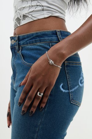 Silver bracelet with heart charm, worn on a hand resting on high-waisted blue denim jeans featuring light blue stitching detail.