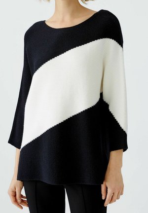 Frau trägt schwarzen Strickpullover mit breitem diagonalen weißen Streifen über Brust und Ärmeln, kombiniert mit schwarzen Hosen.