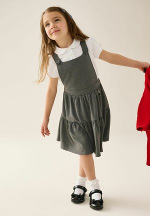 Junges Mädchen mit langen Haaren, das ein graues Kleid mit Trägern, ein weißes Hemd, weiße Socken und schwarze Schuhe trägt und ein rotes Kleidungsstück in der rechten Hand hält.