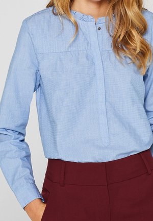 Femme portant un chemisier bleu clair à manches longues avec un col à volants et un pantalon bordeaux, main dans la poche, sur un fond uni.
