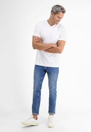 Man with gray hair wearing a white V-neck t-shirt, blue rolled-up jeans, and white sneakers, standing with arms crossed and smiling.