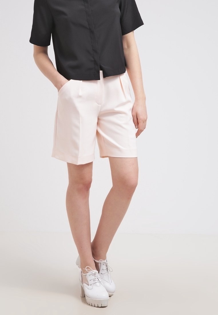 Lichtroze op maat gemaakte shorts met een ontspannen pasvorm, met voorplooien en zijzakken. Gecombineerd met een zwart shirt met korte mouwen en witte schoenen.