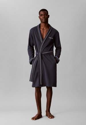 Homme debout pieds nus portant un peignoir gris foncé avec des passepoils blancs et des poches, noué à la taille sur un fond gris uni.