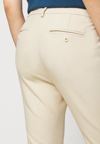 Pantalon beige à coupe ajustée, avec deux poches arrière à boutons, texture lisse et détails cousus.