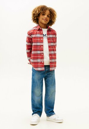 Dziecko o kręconych włosach, ubrane w czerwoną koszulę w kratę na białym swetrze, niebieskie dżinsy i białe trampki, stoi z rękami za plecami.