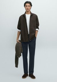 Hnědá košile na knoflíky přes bílé tričko, námořnické kalhoty a tmavě hnědé boty. Drží strukturovaný hnědý svetr. Neutrální pozadí.