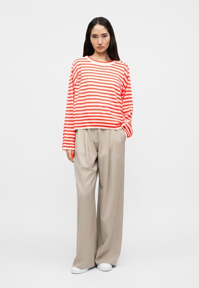 Femme debout portant un chemisier à manches longues rayé rouge et blanc, un pantalon ample beige et des baskets blanches sur un fond blanc uni.