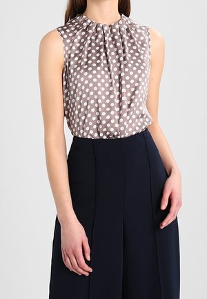 Blouse sans manches à pois blancs sur tissu taupe rentrée dans un pantalon taille haute bleu marine foncé, portée par une personne debout.