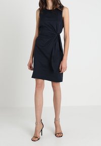 Granatowa sukienka bez rękawów o dopasowanym kroju, z węzłem w talii i gładkiego materiału. W połączeniu z czarnymi sandałami na obcasie.