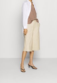 Shorts beige à jambes larges associés à un haut sans manches à motifs dans des tons chauds, et une longue chemise blanche, complétés par des tongs noires.