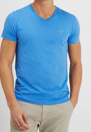 Man wearing a fitted blue V-neck T-shirt and beige pants with one hand in pocket against a plain background.