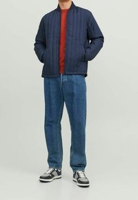 Mörkblå quiltad bomberjacka med ribbad krage, i kombination med en röd topp och blå jeans. Svarta och vita sneakers avslutar outfiten.