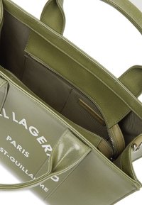 Πράσινη τσάντα ώμου με γυαλιστερή επιφάνεια, ευρύχωρο εσωτερικό και θήκη με φερμουάρ. Εμφανίζει κείμενο λογότυπου στο μπροστινό μέρος.