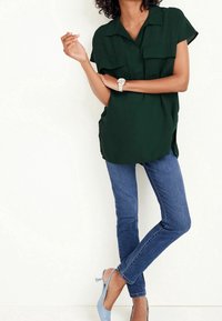 Blouse à manches courtes vert foncé avec deux poches poitrine, associée à un jean skinny bleu et des escarpins slingback bleu clair. Tissu lisse et coupe décontractée.