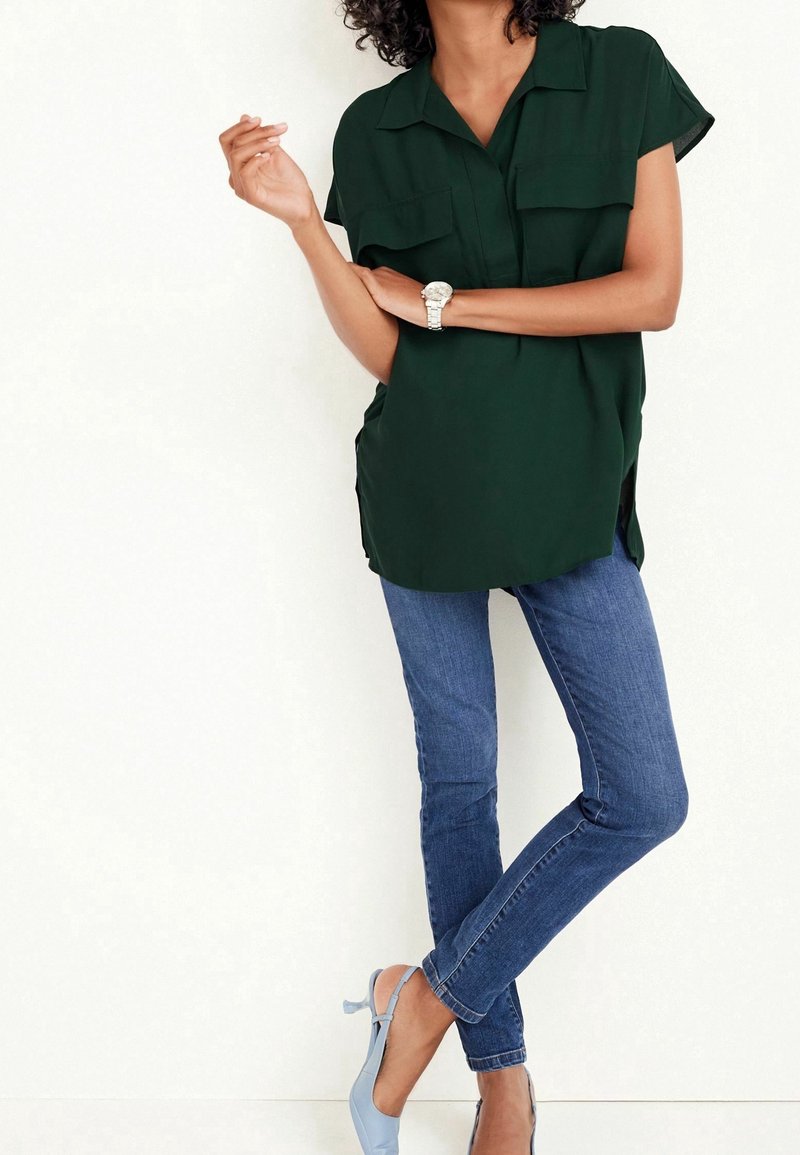 Blouse à manches courtes vert foncé avec deux poches poitrine, associée à un jean skinny bleu et des escarpins slingback bleu clair. Tissu lisse et coupe décontractée.