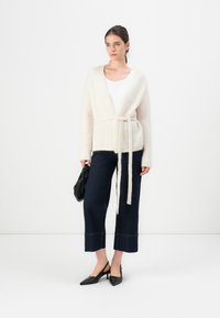 Kvinne med en kremfarget belted cardigan, hvit topp, mørke avkorte jeans, svarte spisse slingback-hæler, holder en svart håndveske, står mot en hvit vegg.