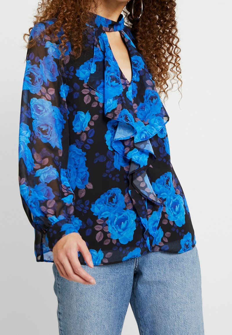 Femme portant un chemisier noir transparent à manches longues avec imprimé floral bleu, devant à volants, encolure goutte d'eau, et jean en denim bleu clair.