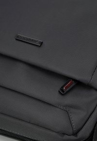 Czarna torba na laptopa wykonana z materiału o teksturowej powierzchni, z zamknięciem na zamek i małą metką z logo. Kompaktowy design z gładkimi krawędziami i wzmocnionymi szwami.