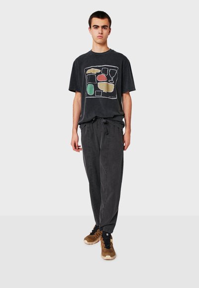 Black graphic t-shirt featuring abstract shapes in yellow, green, red; paired with dark gray tapered pants and brown athletic shoes.