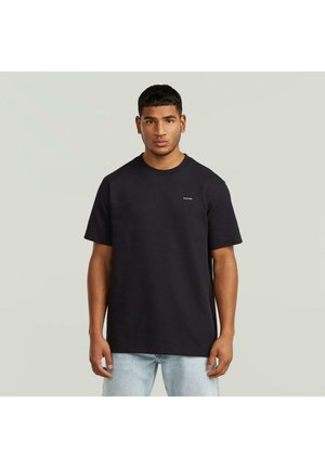 Młody mężczyzna ubrany w gładką czarną koszulkę z krótkim rękawem i jasne jeansy, stojący na tle jasno-szarego tła.