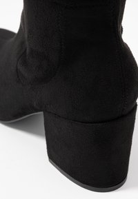 Bottine en daim noir avec talon bloc sur fond blanc, gros plan sur le talon et l'arrière.