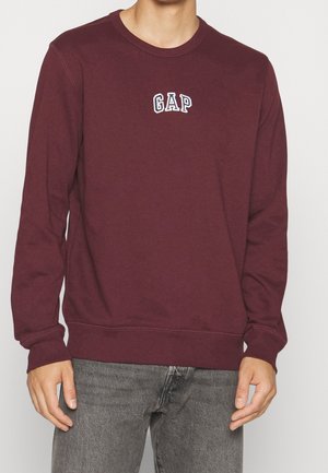 Mann trägt einen dunkelroten Langarm-Sweatshirt mit GAP-Logo auf der Brust und ausgewaschene schwarze Jeans, steht vor einem schlichten hellen Hintergrund.