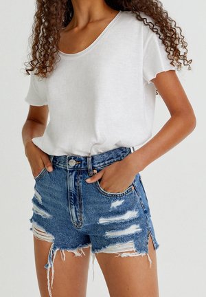 Woman wearing white scoop neck T-shirt tucked into high-waisted distressed blue denim shorts with frayed hems and hands in pockets.