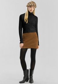Frau mit Leoprint-Beret, schwarzem Rollkragenpullover und Strickjacke, braunem Wildleder-Minirock, schwarzen Strumpfhosen und schwarzen Stiefeletten, aufrecht stehend.