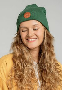 Zielona dziana czapka z prążkowaną teksturą i okrągłą, brązową skórzaną łatką z przodu. Modelka ma długie, falowane włosy i nosi żółty kardigan.