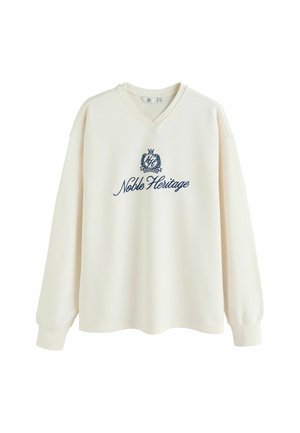 Cream-colored sweatshirt with a V-neck, long sleeves, and "Noble Heritage" embroidered in navy. Soft fabric with a relaxed fit.
